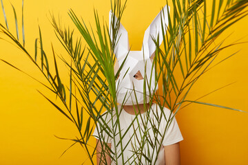 Indoor shot of anonymous woman wearing white T-shirt and paper rabbit mask standing isolated over yellow background, hiding behind green palm leaves, feels scared.
