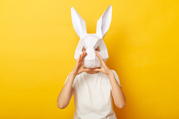 Portrait of woman wearing white T-shirt and paper rabbit mask yelling loud, proclaiming, announcing news or advertisement, standing isolated over yellow background.