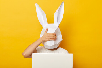 Indoor shot of woman wearing white T-shirt and paper rabbit mask standing isolated over yellow background and working on computer, covering eyes with palm, doesn't want to see something shameful.