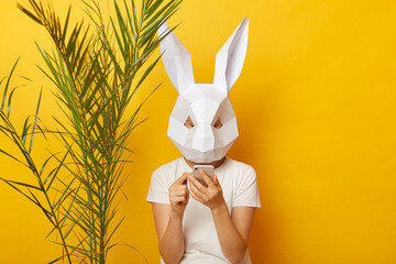Portrait of woman wearing white T-shirt and paper rabbit mask standing isolated over yellow background near green palm leave, using cell phone, chatting, texting with friends.