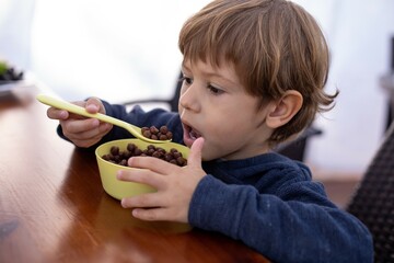 Hungry little boy sitting at wooden table in kitchen and having breakfast with dry chocolate balls closeup. Kid of kindergarten age holding spoon with open mouth eat food. Morning routine, breakfast