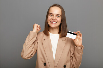 Fototapeta premium Image of happy positive attractive woman with brown hair wearing beige jacket posing isolated over gray background, winning lottery, showing her banking card, clenched fist, celebrating.