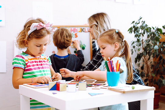Cute little girls with interest fold geometric figures on desk in playroom. Educator with boy learn letters and numbers on magnetic board on background. Preschool, study, daycare centre