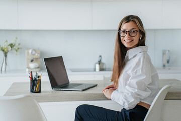 Obraz na płótnie Canvas Beautiful brunette Italian woman in white shirt sits at desk with laptop looks at camera broad smiles works at home office. Successful teacher prepared for distant lesson via internet. Businesswoman