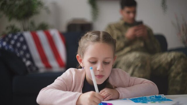 Little girl drawing, military father sitting on couch with gadget, US flag