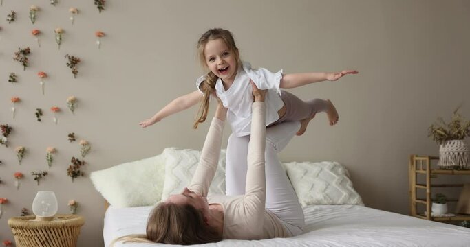 Young woman lying on bed lifts her cute little 4s daughter on arms feel happy, family spend leisure at modern house. Preschooler girl spread arms like plane wings flying in air playing with loving mom