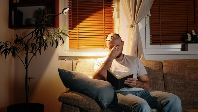 Fatigue and pain in eyes. Bearded Caucasian man wearing eyeglasses and rubbing his eyes is sitting on sofa in cozy evening room and reading book. Concept of education and leisure.