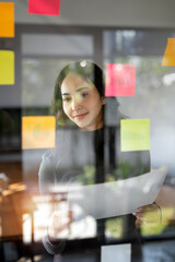 Young smiley attractive businesswoman using sticky notes in glass wall to writing strategy business plan to development grow to success