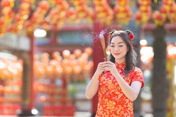 Asian woman in red cheongsam qipao dress is offering incense to the ancestral god inside Chinese...