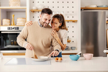 A tender man is preparing eggs for breakfast in the kitchen while his wife is hugging him.