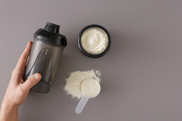 Sports nutrition concept. Top view photo of shaker in hand, whey protein jar and scoop on grey background with copy space. Sport food supplement and fitness diet.