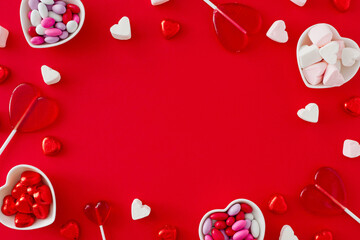 Valentines Day concept. Top view photo of heart shaped lollipops, saucers with chocolate candies and hearts marshmallow on red background with copy space in the middle.