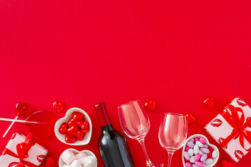 Valentines Day celebration concept. Top view photo of red gift boxes, wine bottle with glasses, heart shaped saucers with candies and lollipops, sprinkles on red background. Flat lay with copy space.