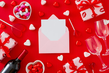 Valentines Day concept. Top view photo of red gift boxes, wine bottle with glasses, heart shaped saucers with candies and marshmallow on red background with paper envelope in the middle.
