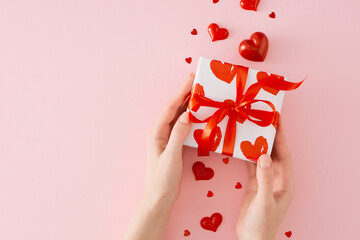 Valentines Day concept. First person top view photo of female hands holding gift box over red heart shaped baubles on pastel pink background with copy space. Love or Valentines holiday card idea.