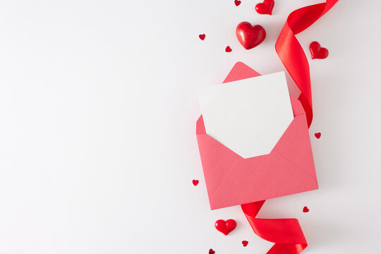 Valentines Day concept. Flat lay photo of envelope with letter, red silk ribbon and heart shaped baubles on white background with copy space. Lovers holiday card idea.