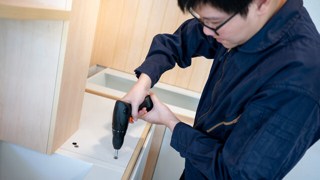 Home renovation or House remodeling concept. Asian male furniture assembler or Interior construction worker man using electric drill screwdriver installing wooden counter and cabinet of new kitchen. - Powered by Adobe