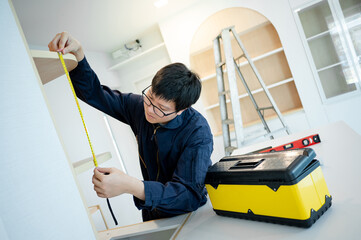 Home renovation or House remodeling concept. Asian male furniture assembler or Interior construction worker man using tape measure installing wooden shelf, cabinet and counter of the new kitchen.