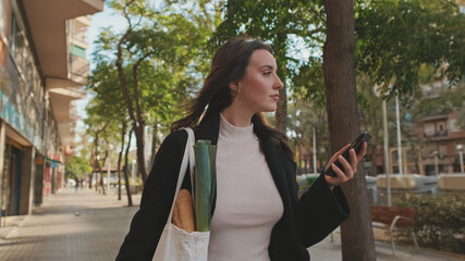Beautiful young woman walking down the street with grocery bag and using mobile phone