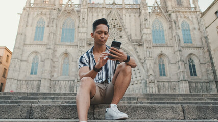 Young man tourist on steps uses mobile phone while sitting on landmarks background