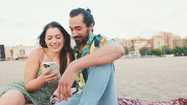 Young couple uses mobile phone while sitting on the beach on buildings background, close-up