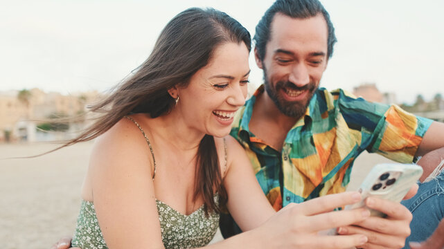 Young couple uses mobile phone while sitting on the beach on buildings background, close-up