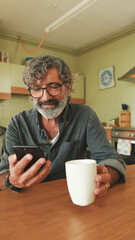 Elderly man drinks coffee from mug and uses mobile phone at home