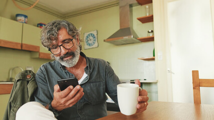 Elderly man drinks coffee from mug and uses mobile phone at home