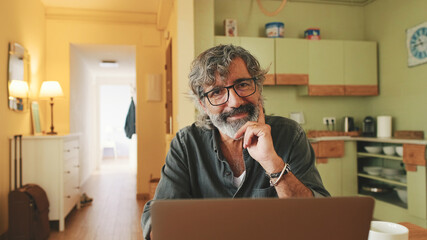 Senior working on laptop sitting in home kitchen and loooks at on camera