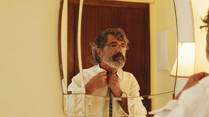 Middle-aged man putting on tie while standing in front of mirror