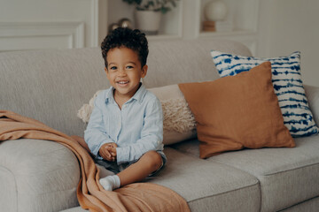 Sweet afro kid boy in casual clothes sitting on comfortable sofa and playing indoors
