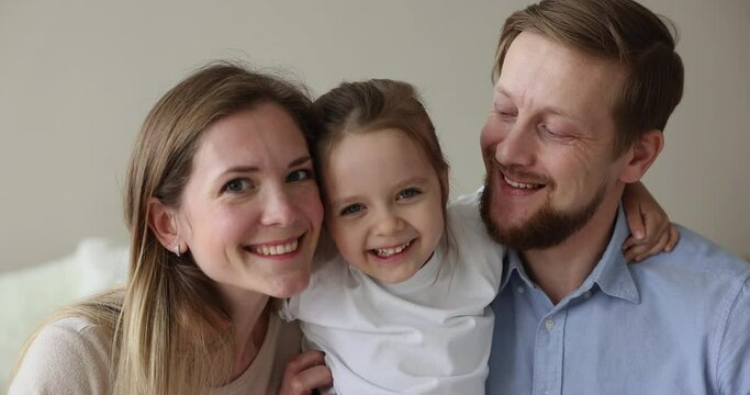 Cheerful young parents and little 4s pretty daughter, close up portrait. Cute preschooler girl hugs their loving mommy and daddy, laughing smile looking at camera. Family ties, happy parenthood, love