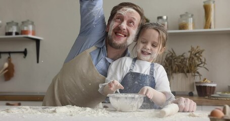 Happy father and cheerful little daughter wear aprons playing together in kitchen, having fun, clap hands and throw flour while make dough for homemade pastry, close up. Family pastime and cooking - Powered by Adobe