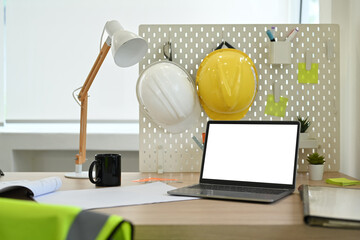 Laptop computer with blank screen, blueprints, hardhat and jacket on wooden table