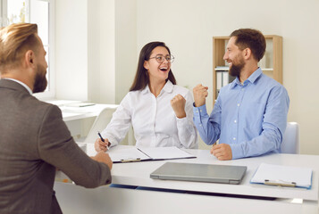 Joyful excited family rejoices during signing of contract of sale or approval of loan. Husband and wife happily clench fists sitting in office in front of bank representative or real estate agent.