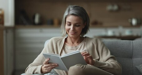 Beautiful middle-aged 45s woman relaxing holds reading book resting on cozy sofa, smile enjoy leisure, carefree weekend with favourite literature and home pastime. Good life habit, development, hobby