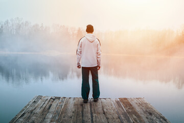 Young man in hoodie, hat and pants standing on wooden pier on pond shore with melancholy fog at...