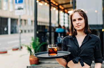 waitress standing in outdoor cafe. girl the waiter holds a tray with a glass of alcoholic cocktail