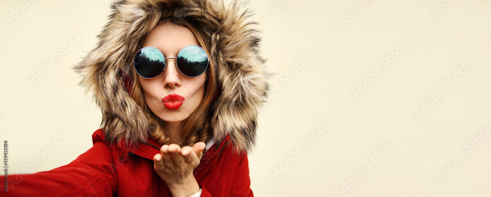 Wall mural Portrait of stylish woman blowing her lips sends air kiss stretching hand for taking selfie with smartphone wearing jacket with fur hood