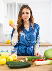 Obraz premium Satisfied woman in dressing gown eating apples in kitchen during breakfast