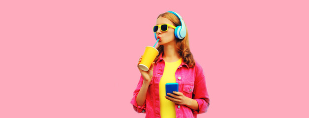 Portrait of stylish young woman listening to music in headphones with cup of fresh juice wearing...