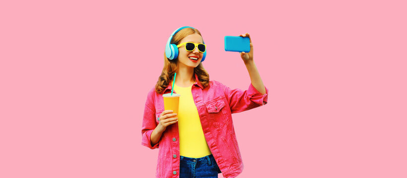 Portrait of happy smiling woman taking selfie with smartphone listening to music in wireless headphones wearing jacket on vivid pink background