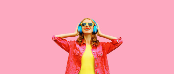 Portrait of happy smiling young woman in headphones listening to music wearing jacket on pink...
