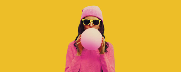 Fashionable portrait of stylish cool young woman inflating chewing gum or balloon wearing pink hat on yellow background, blank copy space for advertising text