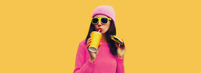 Portrait of stylish young woman drinking juice with burger fast food on yellow colorful background