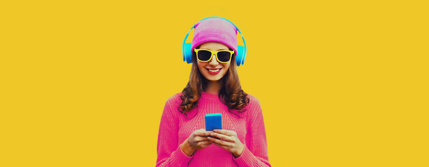 Portrait of modern young woman in wireless headphones listening to music with smartphone wearing knitted sweater, pink hat on yellow background - obrazy, fototapety, plakaty