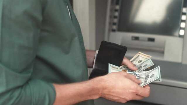 Close-up view of hands of adult young man receiving money from ATM. Modern male withdrawing dollar bills counting putting in wallet. Making currency financial operations and transactions.