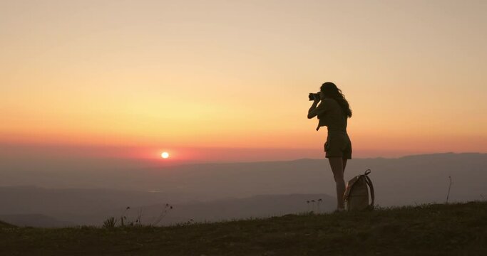 Millennial woman photographer stands at orange summer sunrise and takes pictures of incredible mountain landscape. Silhouette of travel photographer woman at dawn in early morning. Woman takes photos.
