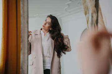 Gorgeous brunette hispanic woman at mirror trying on beige coat at fitting room. Attractive calm caucasian model with long loose hair prepares for date. Women beauty. Hairstyle. Businesswoman at home.