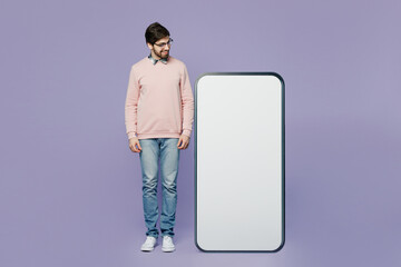 Full body smiling happy young IT man he wear casual clothes pink sweater glasses look at big huge blank screen mobile cell phone smartphone with area isolated on plain pastel light purple background.